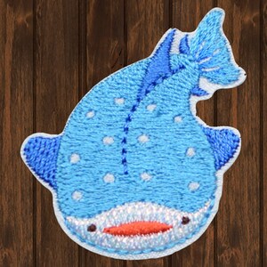 May include: A blue and white embroidered patch of a whale shark with a white background. The whale shark has a light blue body with white spots and a dark blue fin.