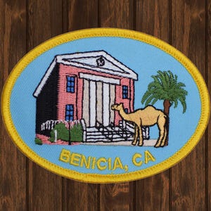 May include: Embroidered patch with a pink building, a camel, and a palm tree. The text "BENICIA, CA" is below the image.