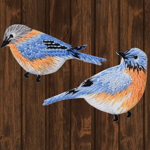 May include: Two embroidered bluebird patches. The birds feature blue, orange, and white thread, with black accents on the wings and feet. The patches are set against a dark brown, wood-grain background.