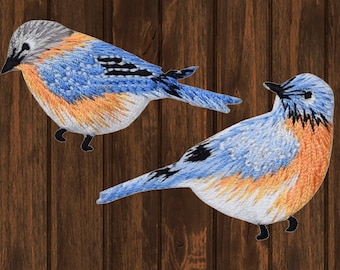 Può includere: Due toppe ricamate di uccelli azzurri. Gli uccelli presentano fili blu, arancioni e bianchi, con accenti neri sulle ali e sulle zampe. Le toppe sono ambientate su uno sfondo marrone scuro con venature del legno.