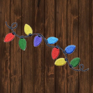 Puede incluir: Cadena bordada de luces navideñas en rojo, verde, amarillo, azul y morado. Las luces están en un cable negro, con un remolino decorativo al final. El fondo es una veta de madera marrón oscuro.