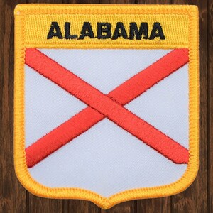 May include: A yellow embroidered patch with a white background and a red cross. The text "ALABAMA" is embroidered in black on the top of the patch.