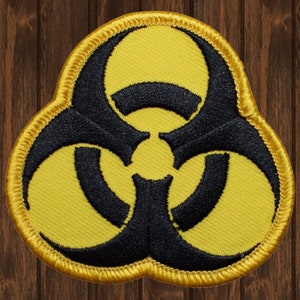 May include: Embroidered patch featuring a black biohazard symbol on a yellow background. The patch has a gold border and a textured appearance. The design is a warning symbol, commonly associated with hazardous materials.