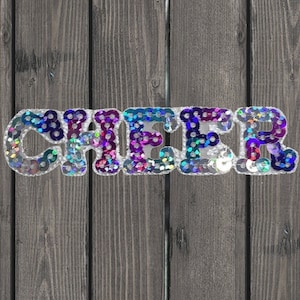 May include: A silver sequined patch with the word "CHEER" in rainbow colors. The patch is on a wooden background.