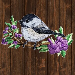 Puede incluir: Parche bordado de un carbonero gris y negro posado en una rama con flores moradas. El pájaro está mirando hacia la izquierda y tiene un pequeño pico negro. Las flores son de un morado claro y tienen hojas verdes.