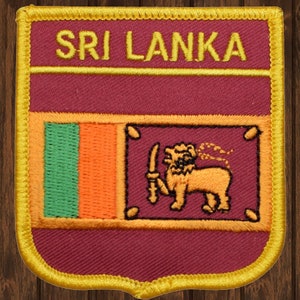 Può includere: Una toppa in tessuto color granata con un bordo dorato e le parole "SRI LANKA" in lettere dorate. La toppa presenta la bandiera dello Sri Lanka con una striscia verde, arancione e gialla a sinistra e un leone con una spada a destra.