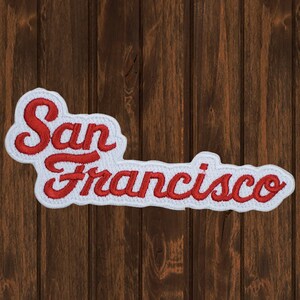 May include: White embroidered patch with red lettering that reads "San Francisco" on a wood background.