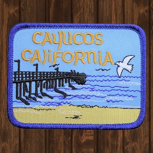May include: Embroidered patch with a blue background, featuring a pier with a white seagull flying above it. The text "Cayucos California" is embroidered in yellow on the patch.