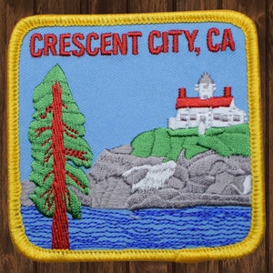 May include: Embroidered patch with a yellow border, depicting a lighthouse on a cliff with a green hillside, a blue body of water, and a green tree with a red trunk. The text "CRESCENT CITY, CA" is at the top of the patch.