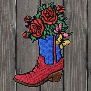 Puede incluir: Parche bordado de una bota vaquera roja con un eje azul lleno de flores rojas, rosas y amarillas. La bota tiene un tacón marrón y una suela marrón.