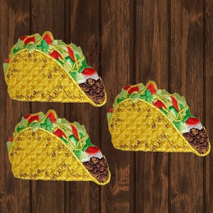 May include: Three embroidered taco patches. Each patch features a yellow tortilla filled with brown meat, red tomatoes, and green lettuce. The patches are on a brown wooden background.
