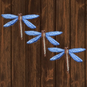 Könnte beinhalten: Drei blaue Libellen-Stickerei-Aufnäher auf braunem Holzgrund.