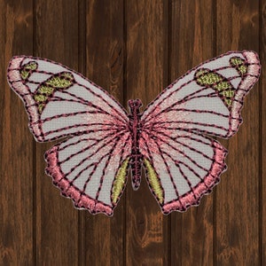 May include: A pink and white embroidered butterfly with gold accents. The butterfly is on a brown wooden background.