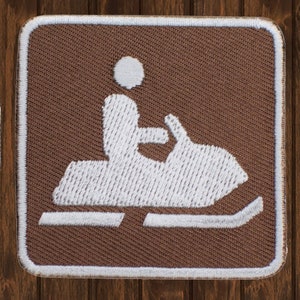 May include: White embroidered snowmobile icon on a brown fabric patch. The snowmobile is outlined in white and has a white figure of a person riding it.