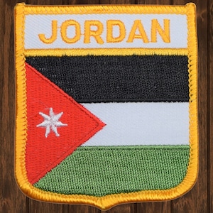May include: An embroidered patch with the flag of Jordan. The patch is yellow with a red, white, black, and green flag in the center. The flag has a seven-pointed star in the red triangle. The words "JORDAN" are embroidered in yellow at the top of the patch.