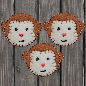 Puede incluir: Tres caras de mono de fieltro con pelaje marrón, caras blancas y ojos negros. Los monos tienen narices rojas y sonrisas cosidas.