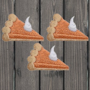 May include: Three embroidered patches of pumpkin pie slices with whipped cream. The pie crust is tan, the filling is orange, and the whipped cream is white. The patches are on a wooden background.
