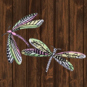 May include: Two embroidered dragonfly patches. The dragonflies feature intricate wing designs with pastel colors, including pink, yellow, and green, set against a white background. The patches are placed on a dark brown wooden surface.
