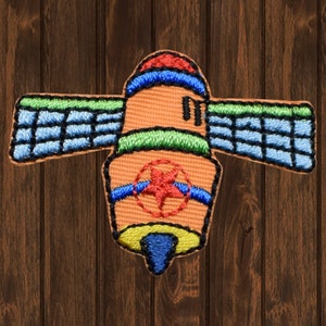 May include: A colourful embroidered patch featuring a satellite with a red star at its centre. The satellite is orange with blue and green details. The patch is ideal for adding a unique touch to clothing, bags, or other accessories.