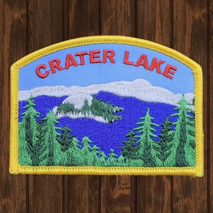 May include: Embroidered patch featuring a scenic view of Crater Lake. The patch has a blue sky, white clouds, and green trees. The words "CRATER LAKE" are in red letters across the top. The patch is bordered with yellow.
