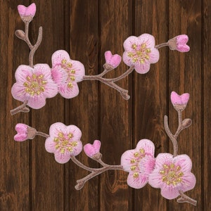 May include: Embroidered pink cherry blossom patches with gold centers and brown stems. The floral patches are arranged on a dark brown wood grain background. The patches feature detailed stitching and a delicate design.