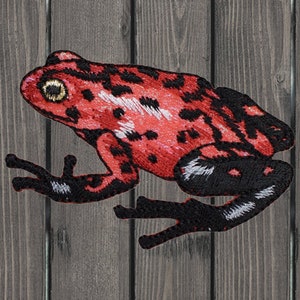 Puede incluir: Parche de rana bordado en rojo y negro. La rana tiene una franja blanca en la espalda y un ojo amarillo.