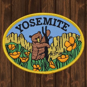 May include: A yellow oval embroidered patch with the word "Yosemite" in black lettering. The patch features a brown bear hugging a tree with orange poppies in the foreground.