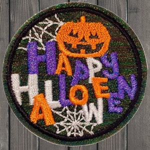 May include: A round, green, embroidered patch with a white, orange, and purple "Happy Halloween" message. The patch features a jack-o'-lantern and spiderwebs.