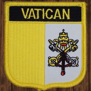 May include: A yellow and white embroidered patch with the word "VATICAN" in black lettering at the top and a white papal coat of arms on a white background.