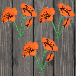 Op de afbeelding: Vijf oranje geborduurde klaproosbloem-patches met groene stelen en bladeren. De patches bevinden zich op een houten achtergrond.