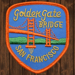 Puede incluir: Un parche bordado azul y naranja con el texto "Golden Gate Bridge San Francisco". El parche presenta un puente rojo con torres y cables de suspensión.