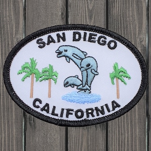 May include: White oval patch with black border, featuring the text "SAN DIEGO CALIFORNIA" in black embroidery. Three blue dolphins jump out of the water, with two palm trees on either side.