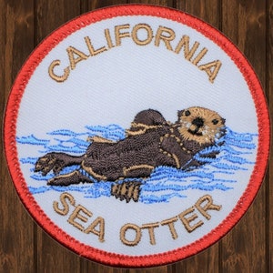 Puede incluir: Parche bordado con una nutria marina marrón nadando en agua azul. El parche es blanco con un borde rojo y el texto "CALIFORNIA SEA OTTER" en marrón.