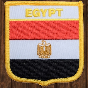 Peut inclure: Un écusson brodé rouge, blanc et noir avec le mot "EGYPT" en lettres jaunes et un emblème d'aigle doré au centre. L'écusson est entouré d'une bordure jaune.