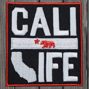 May include: A black and white embroidered patch with the text "CALI LIFE" in white. The patch features a red bear and a star on a white stripe, representing the California flag. The patch is outlined in red.