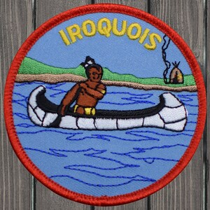 Peut inclure: Un écusson rond bordé de rouge avec un fond bleu. L'écusson représente un Amérindien pagayant dans un canoë sur un lac bleu. Le texte "IROQUOIS" est écrit en jaune au-dessus du canoë. Un petit tipi brun avec de la fumée qui sort du sommet se trouve en arrière-plan.