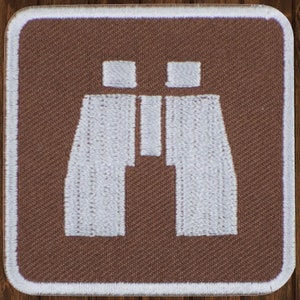 May include: A brown fabric patch with a white embroidered image of binoculars. The patch is square shaped with white stitching around the edges.