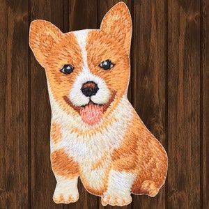 May include: Embroidered patch of a brown and white corgi puppy with a pink tongue sticking out. The puppy is sitting with its front paws crossed and its back legs extended.