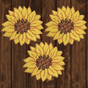 Puede incluir: Tres parches bordados de girasol en amarillo y marrón. Los girasoles están dispuestos en forma triangular sobre un fondo de madera marrón.