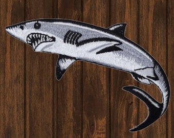 May include: A white and black embroidered shark patch. The shark is swimming with its tail curled up. The patch is on a wood background.