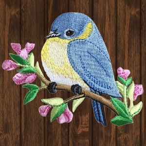 May include: Embroidered patch of a bluebird perched on a branch with pink flowers. The bird has a yellow breast and a blue head and back. The flowers are pink with green leaves.