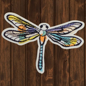 May include: Embroidered dragonfly patch with a white border. The dragonfly features wings with yellow, orange, blue, and purple accents. The body is a mix of orange and blue. The patch is set against a brown wooden background.