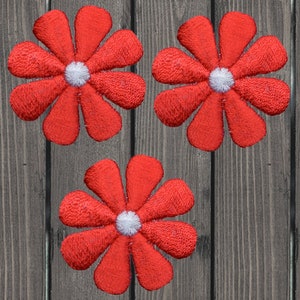 May include: Three embroidered red daisy flower patches with white centers. The flowers have six petals each and are arranged on a wooden background. These patches could be used for clothing or craft projects.