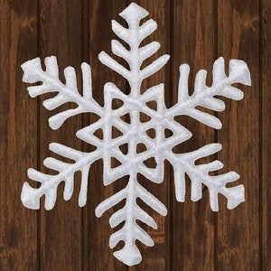 Könnte beinhalten: Ein weißes Schneeflocken-Ornament mit detaillierten Mustern, vor einem dunkelbraunen Holz-Hintergrund. Die Schneeflocke hat sechs symmetrische Spitzen und eine strukturierte Oberfläche, passend für Winter- oder Festtagsdekorationen.