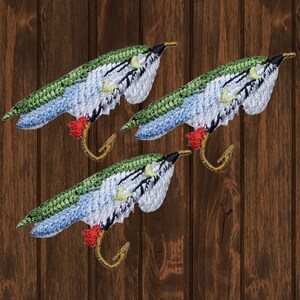 Könnte beinhalten: Drei gestickte Angelköder-Patches mit grünem, blauem, weißem, rotem und goldenem Garn. Die Köder befinden sich auf einem braunen Holzgrund.