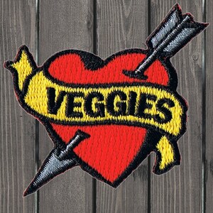 Op de afbeelding: Een rood hart met een gele banner waarop "VEGGIES" staat en dat door een zilveren pijl wordt doorboord. Het hart is geborduurd en heeft een zwarte omtrek.