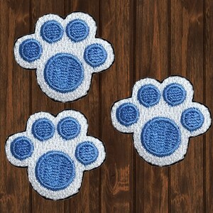 Può includere: Tre toppe termoadesive a forma di impronta di zampa blu e bianca. Le impronte di zampa sono ricamate con filo blu su uno sfondo bianco.