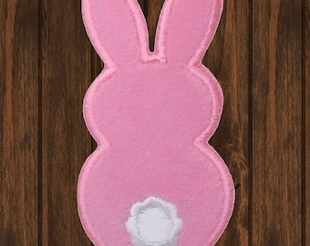 May include: A pink bunny-shaped patch with a white fluffy tail. The patch is outlined with a light pink border. The background is a wooden surface.