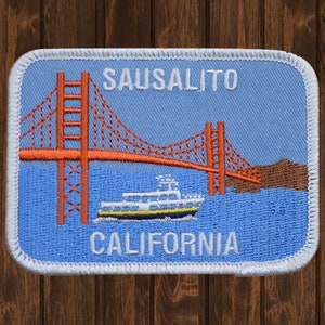Op de afbeelding: Een blauwe en witte geborduurde patch met de tekst "SAUSALITO CALIFORNIË" en een afbeelding van de Golden Gate Bridge en een veerboot.