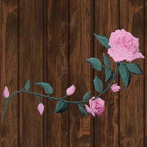 May include: A pink embroidered rose with green leaves and buds on a brown wooden background.